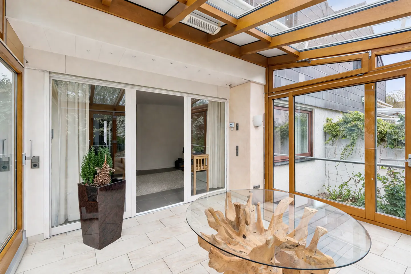 Sunroom with glass roof, tile floor, and glass table with a driftwood base. Sliding glass doors lead to a patio with greenery.
