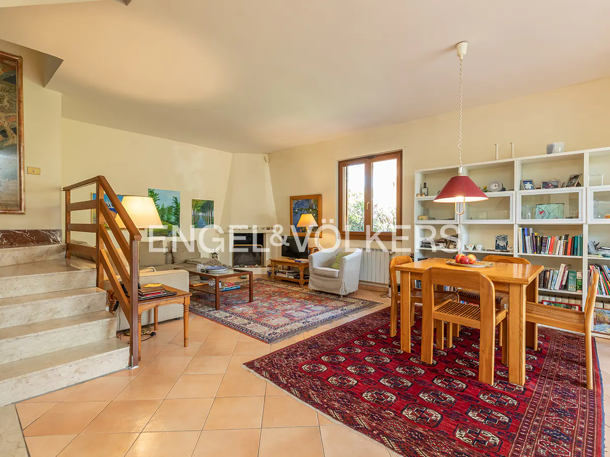 A bright living room with a dining table, chairs, and a bookshelf. A staircase is on the left, and a fireplace is in the background.