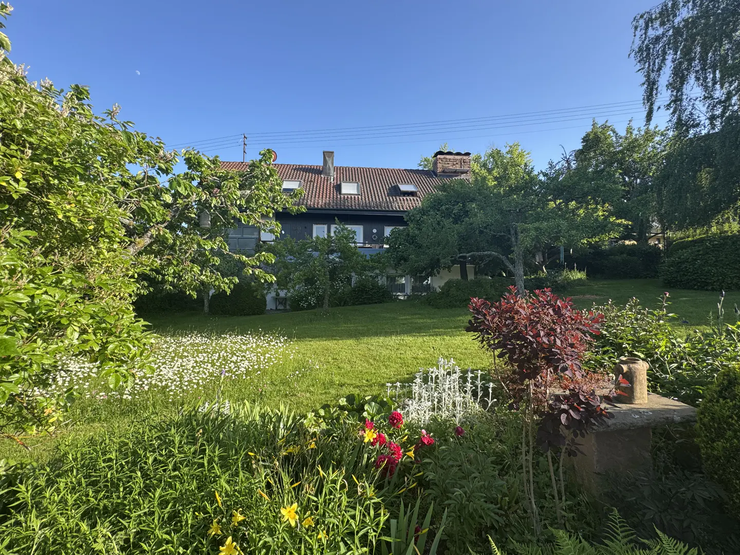 A two-story house with a tiled roof sits behind a lush green garden filled with flowers, trees, and a stone pedestal.