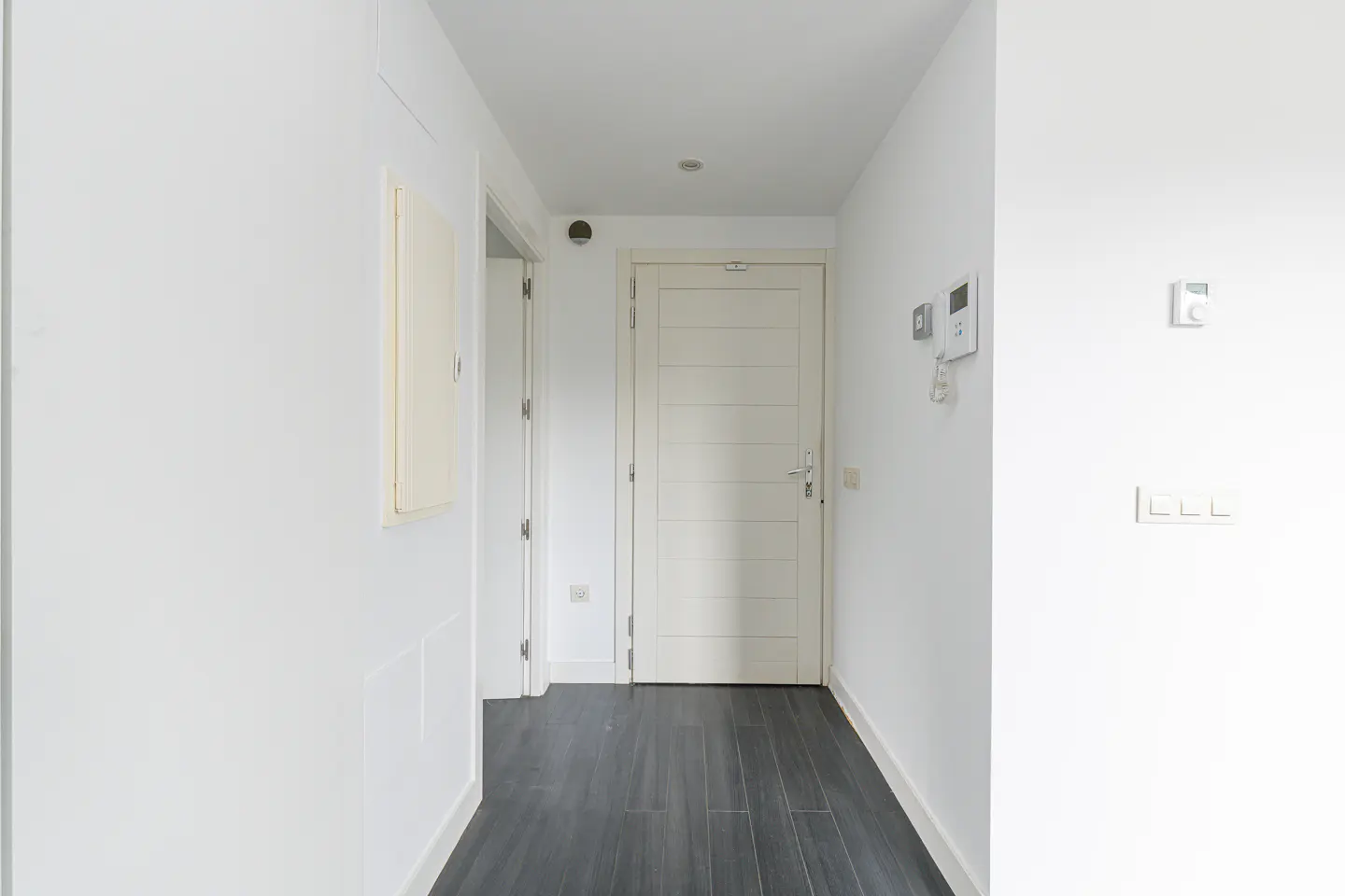 A bright, narrow hallway with white walls and dark wood floors. A white door is at the end of the hall.