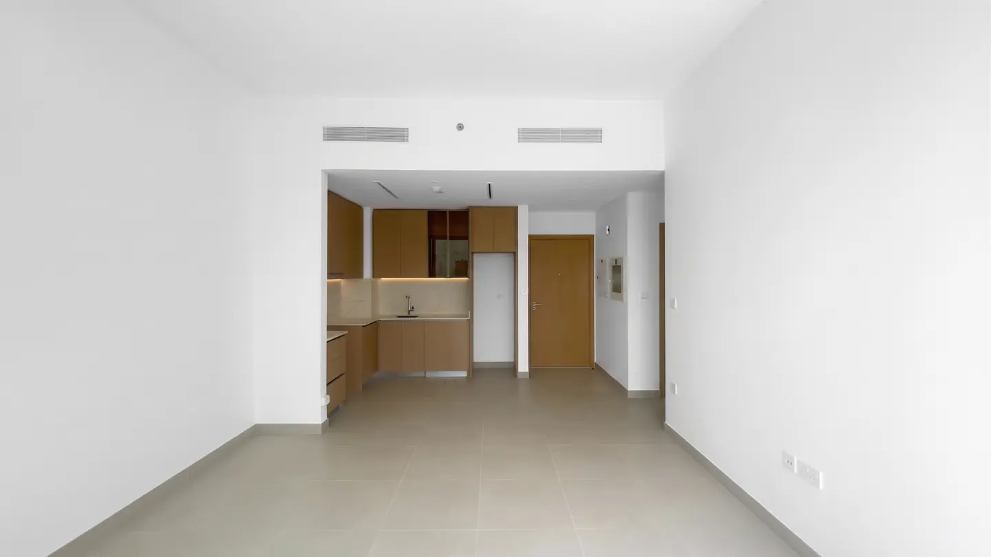 An empty apartment with white walls and beige tile floors. A kitchen with light brown cabinets is visible in the background. A brown door is on the right.