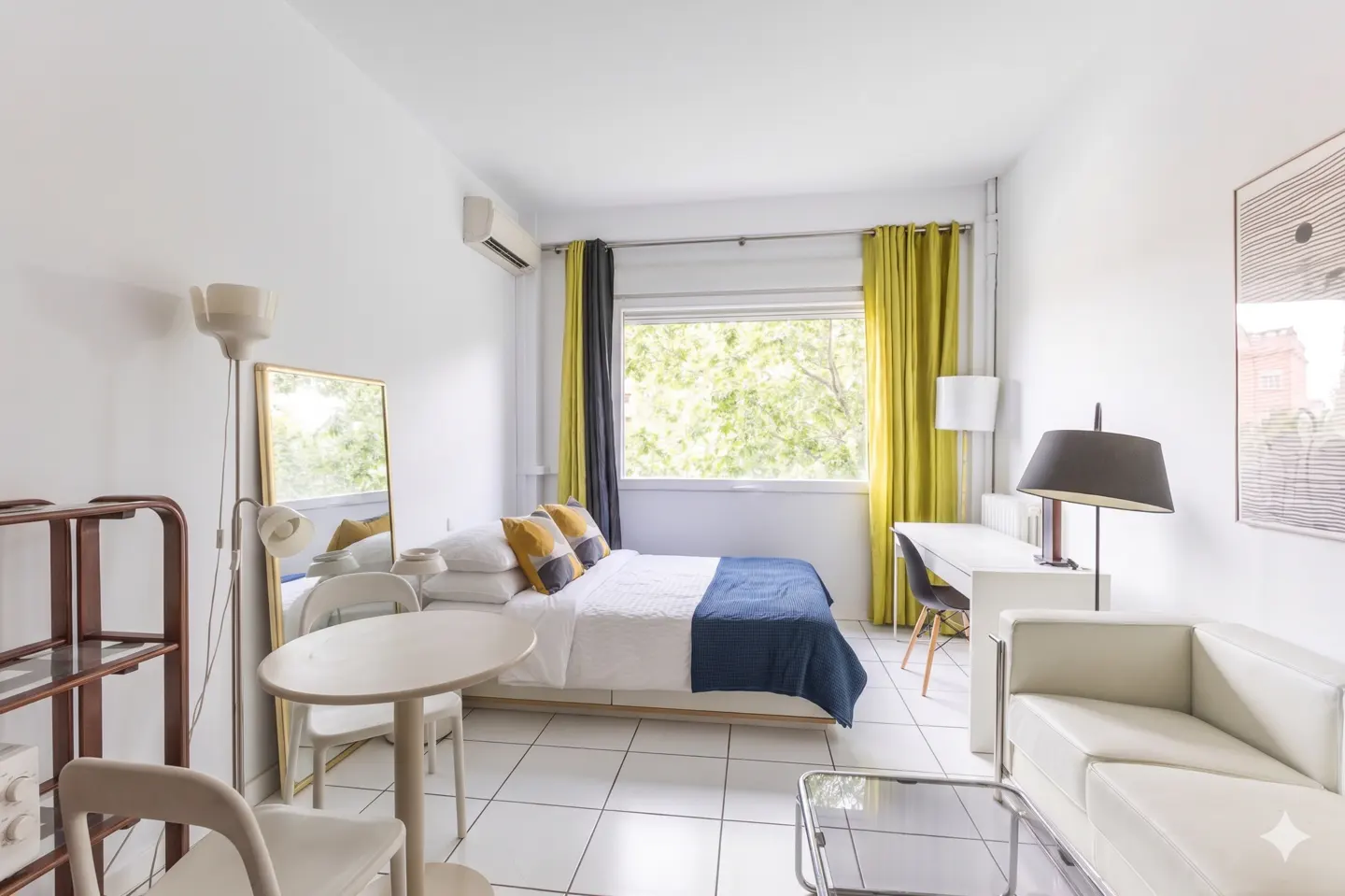 Bright studio apartment with white walls, tile floors, and a bed with yellow pillows and a blue blanket. A desk, sofa, and table are also present.