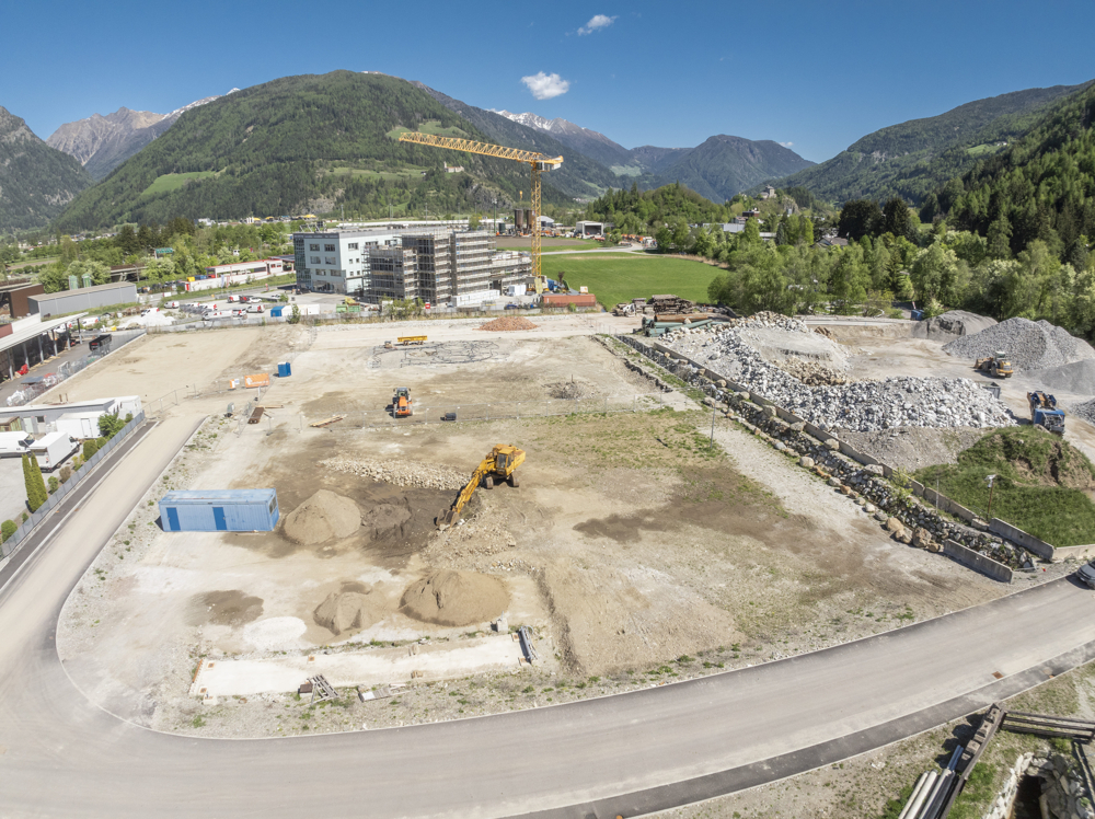 Aerial view of a construction site with heavy machinery, piles of dirt, and a yellow crane against a mountain backdrop. [AI-generated text]