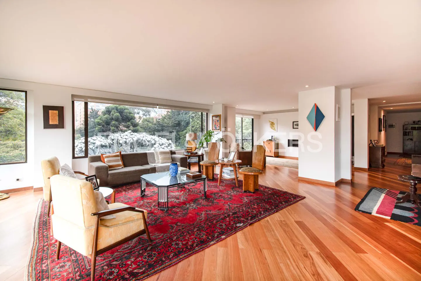 Bright living room with hardwood floors, a red rug, and a gray sofa. Large windows offer a view of lush greenery.