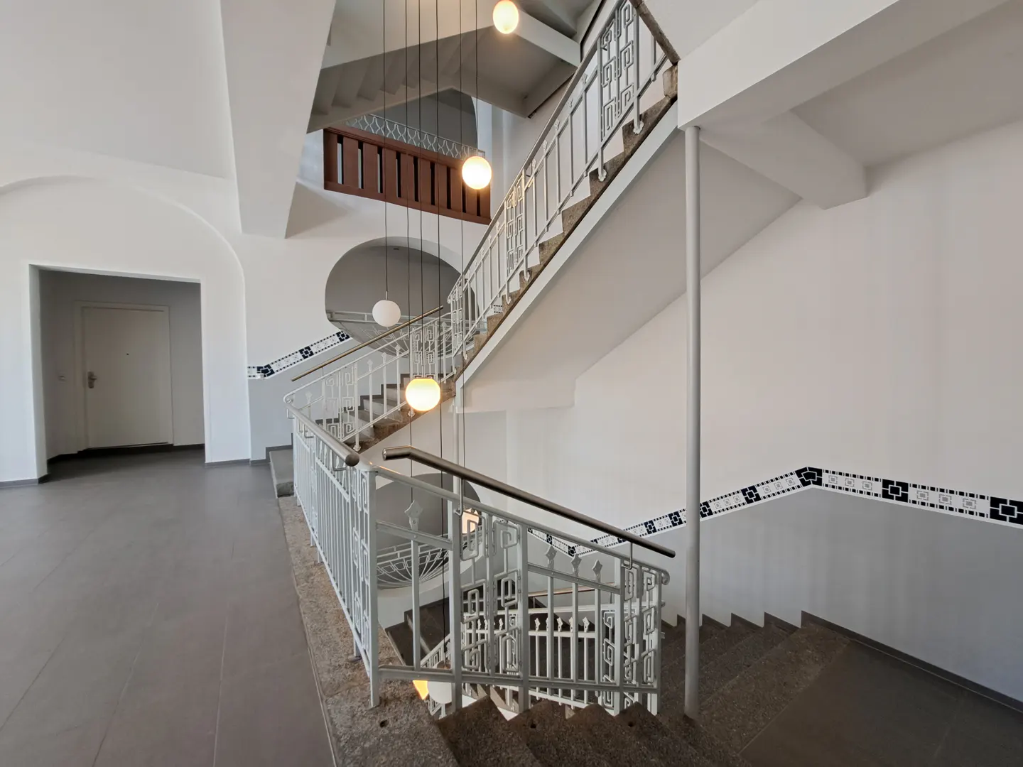 Interior view of a white stairwell with gray stairs and white metal railings. Three globe lights hang from the ceiling.