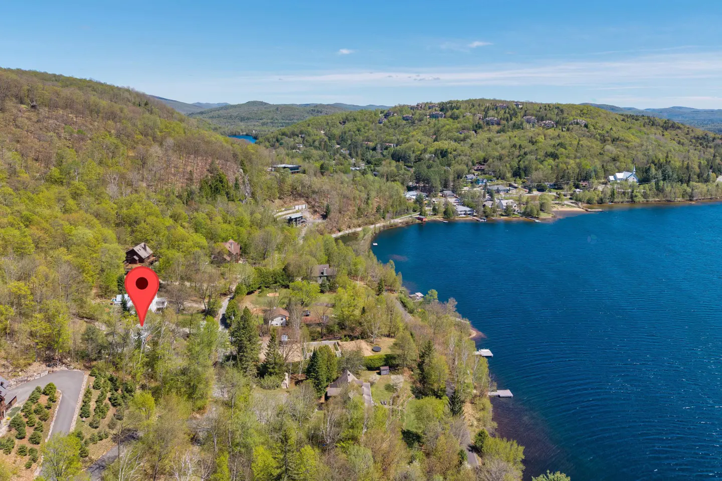 Aerial view of a lakeside property with a red map pin, surrounded by green trees and blue water under a clear sky.