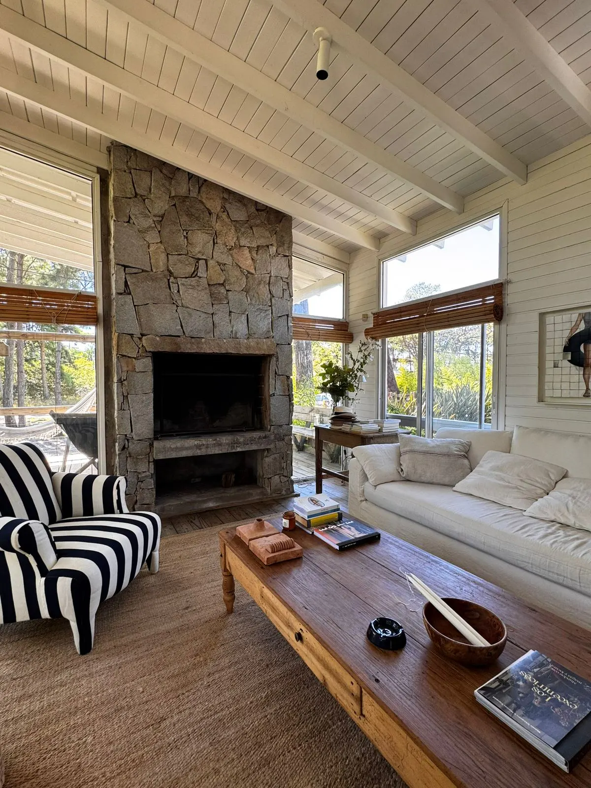 Bright living room with stone fireplace, white sofa, and black and white striped chair. Large windows offer views of greenery.