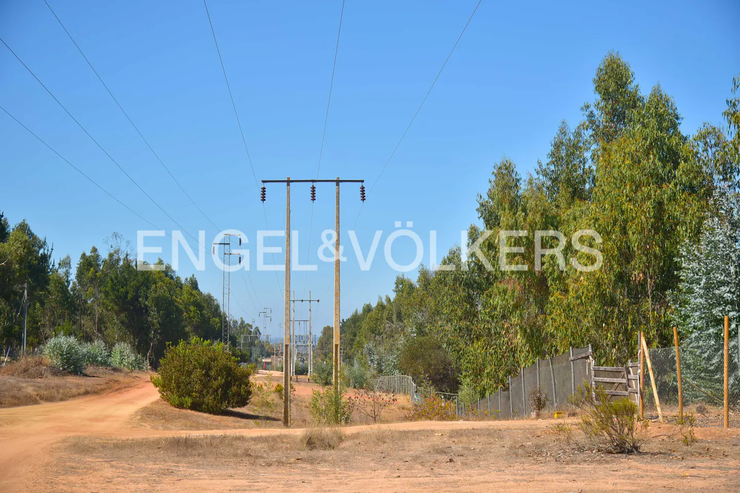 Dirt road with power lines and green trees under a blue sky. Engel & Völkers logo overlayed.
