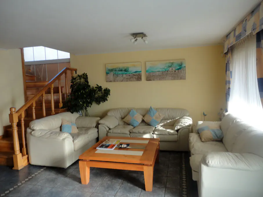 Living room with three white sofas, blue pillows, a wooden coffee table, and a staircase in the background.