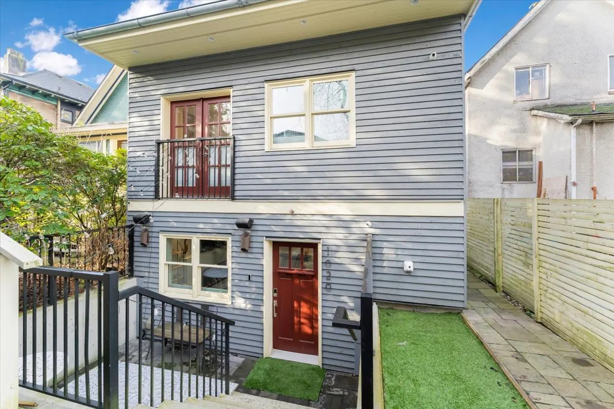Exterior view of a two-story gray house with a red front door and a small balcony with red doors. A staircase leads to the entrance.
