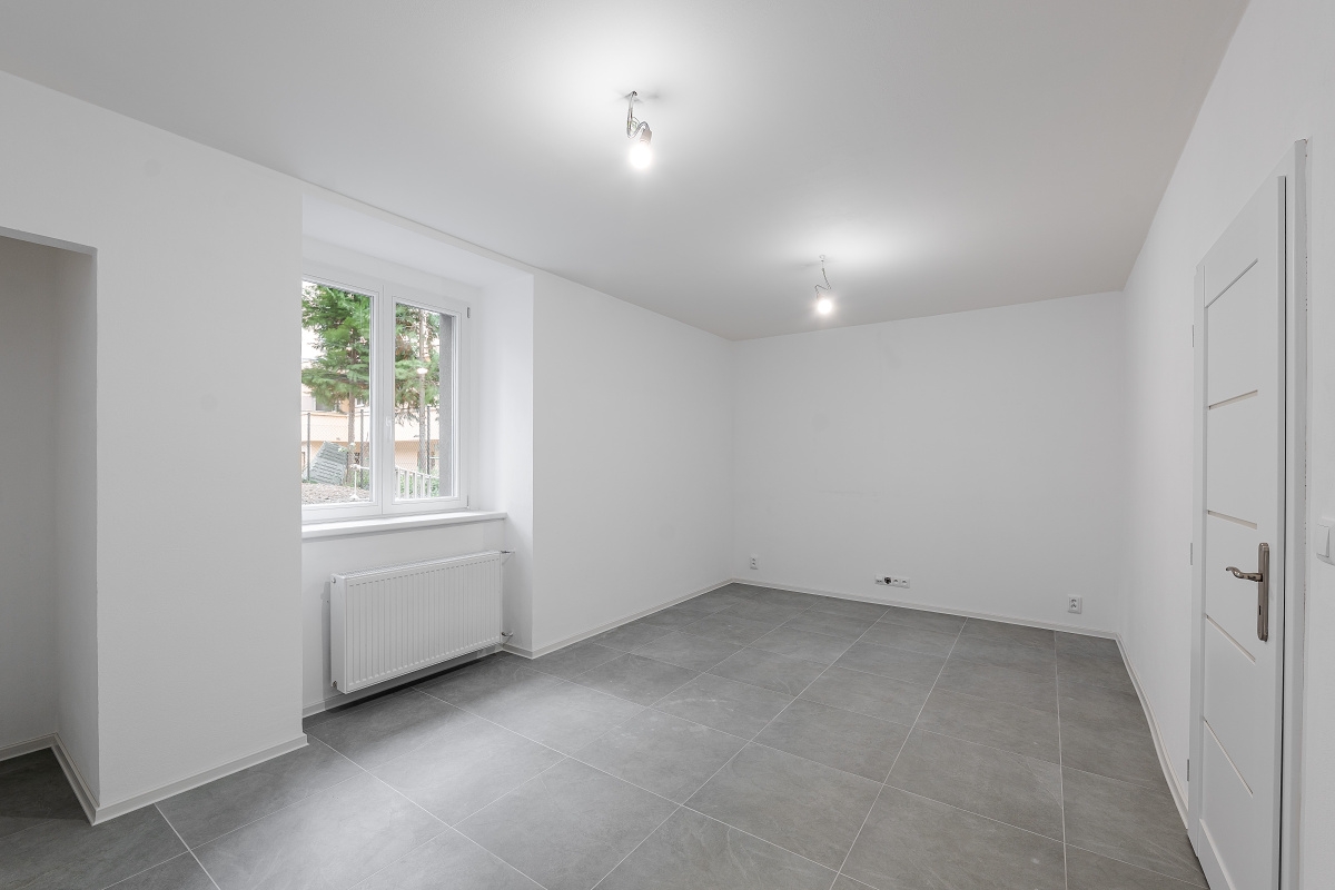 Bright, empty room with white walls, gray tile floor, and a window showing trees outside. A white door is on the right.