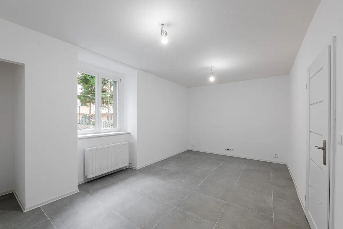 Bright, empty room with white walls, gray tile floor, and a window showing trees outside. A white door is on the right.