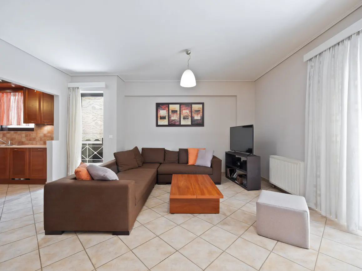 Living room with brown sectional sofa, wood coffee table, TV, and tile floor. Kitchen visible in background.