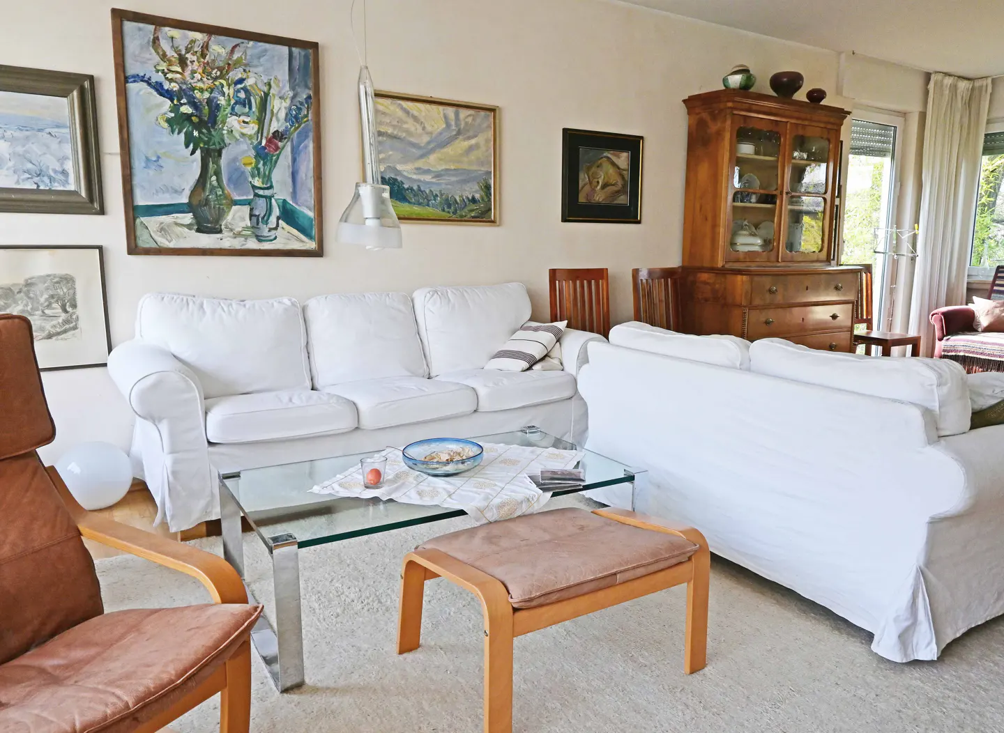 A bright living room with white sofas, a glass table, and wooden furniture. Artwork adorns the walls.
