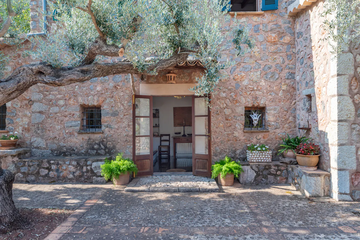 Stone house with open double doors, potted plants, and an olive tree. Interior view shows a table and chairs.