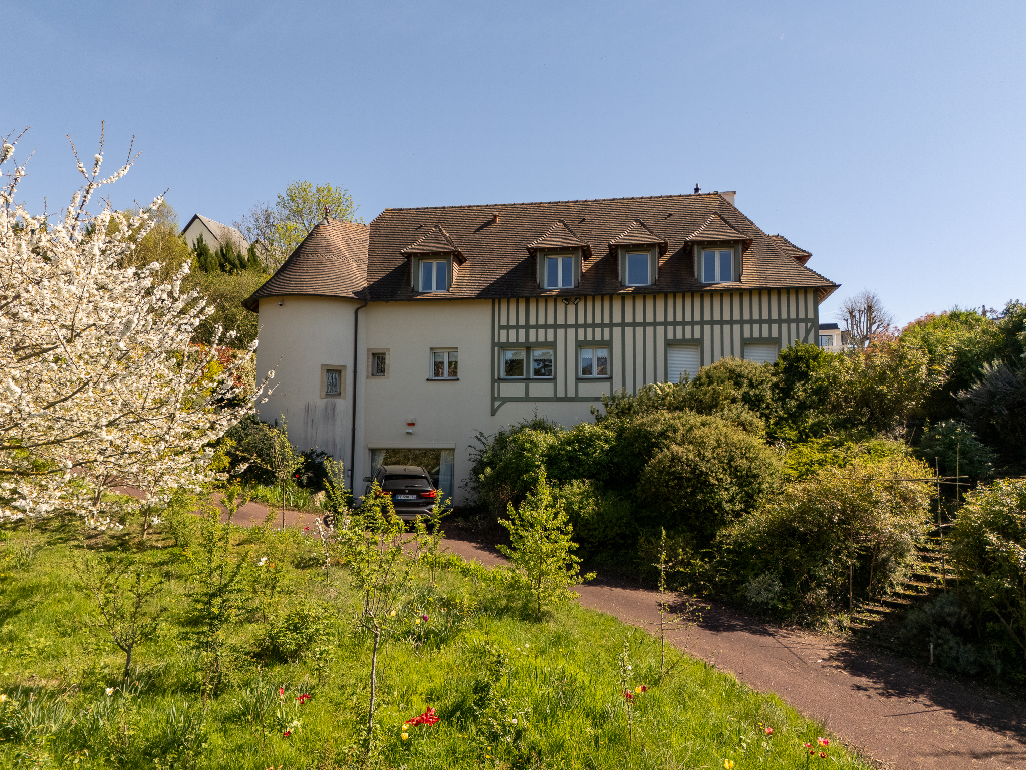 Exterior view of a two-story house with a brown roof, white walls, and green trim, surrounded by lush greenery and a blooming tree. A car is parked in the garage.