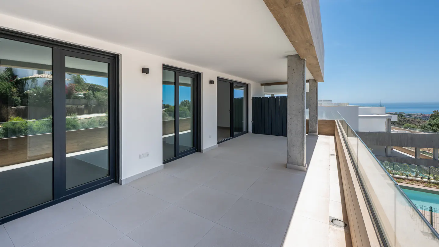 A bright, modern balcony with gray tile flooring, black-framed sliding glass doors, and a glass railing overlooking a scenic view.