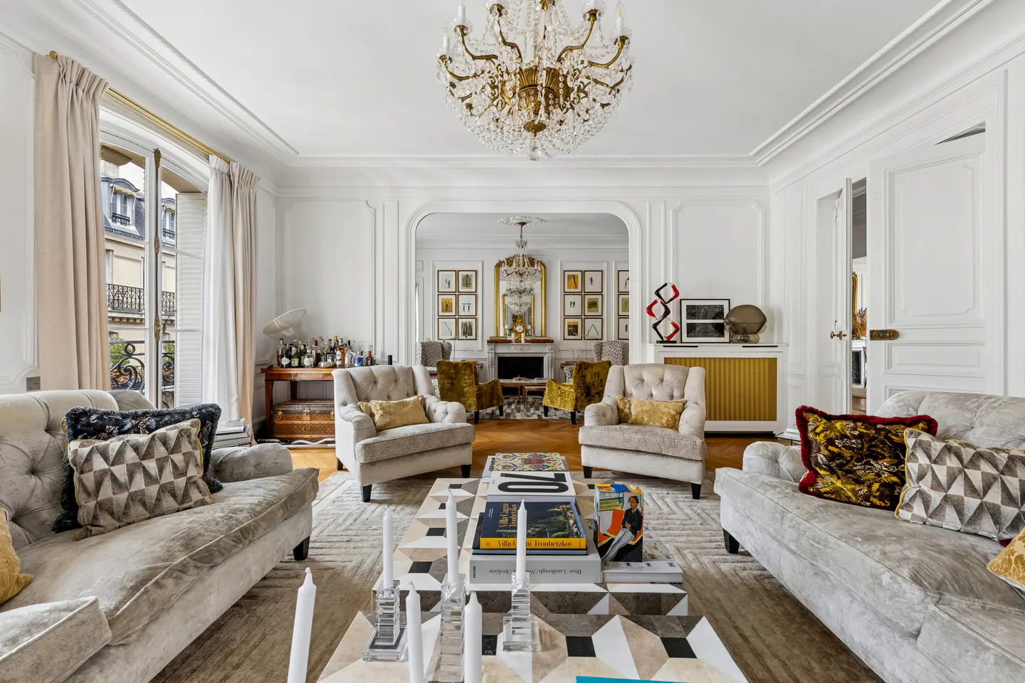Elegant living room with white walls, two sofas, armchairs, and a geometric coffee table. A crystal chandelier hangs from the ceiling.