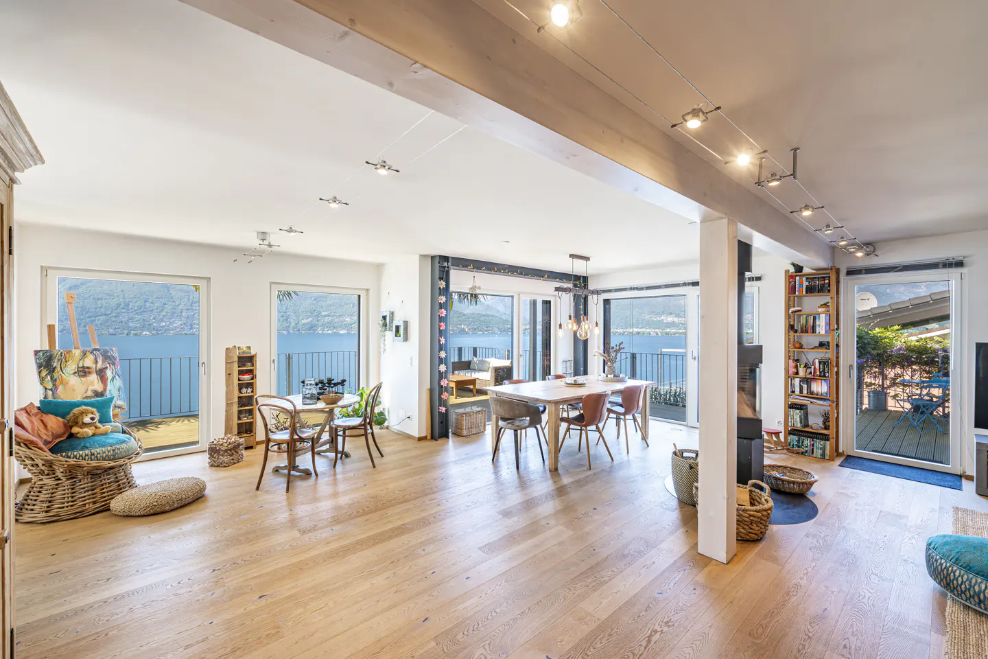 Bright, open-plan living space with wood floors, dining table, and lake views through large windows. Artwork and seating add color.
