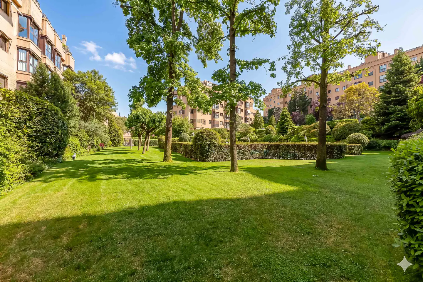 Lush green lawn with trees in front of tan apartment buildings under a blue sky.