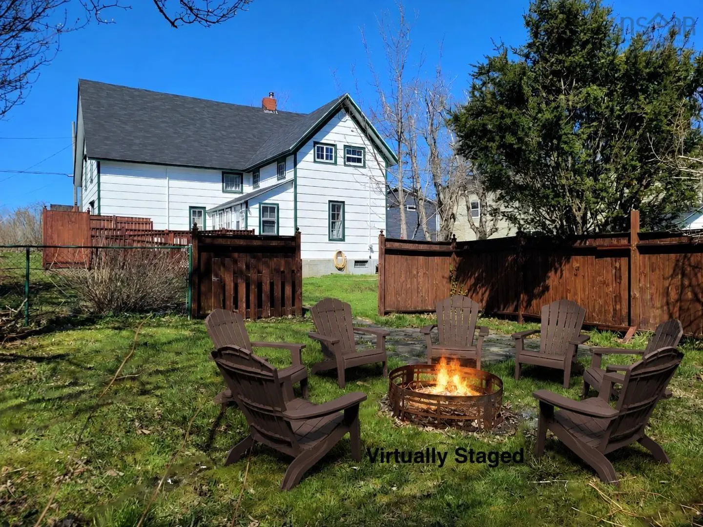 Backyard with a fire pit surrounded by six brown Adirondack chairs on green grass, a white house with a black roof in the background.