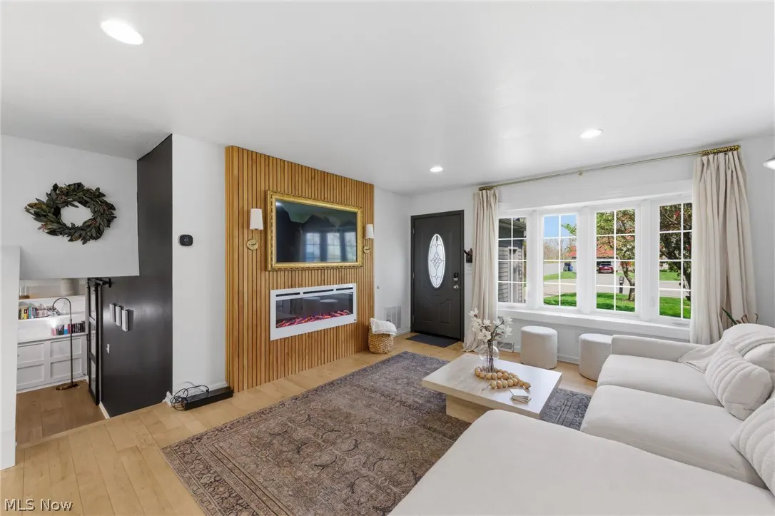 Bright living room with a white sectional sofa, a coffee table, and a fireplace under a TV on a wood-paneled wall. A large window and a black front door are visible.