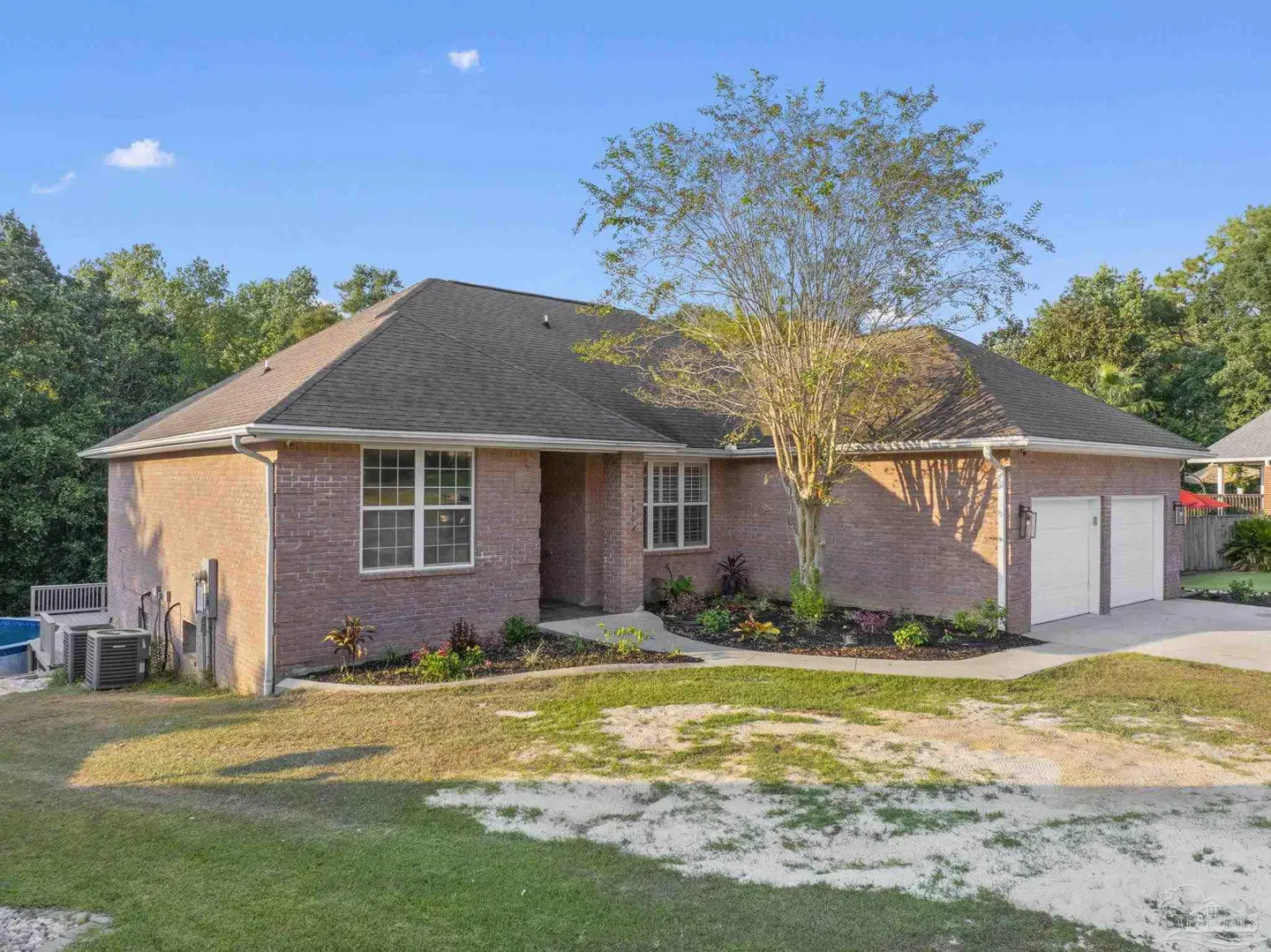 A single-story red brick house with a gray roof, white garage doors, and a tree in the front yard.
