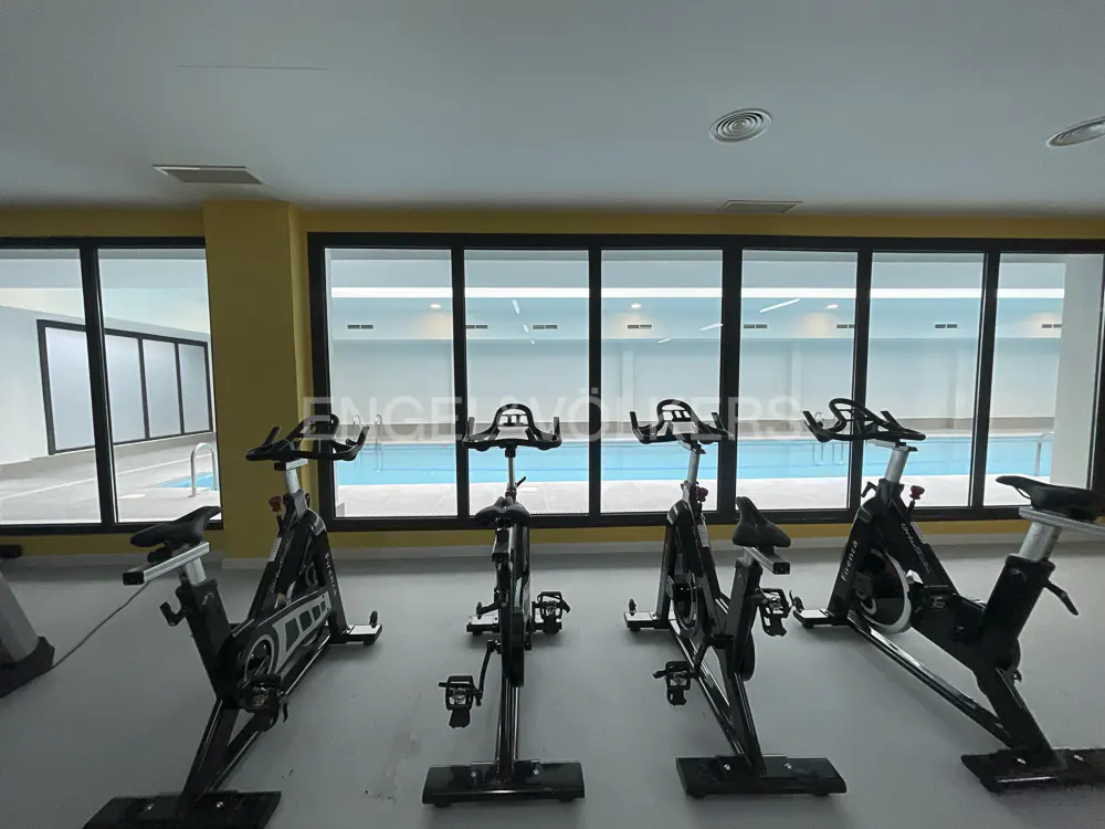 Indoor gym with stationary bikes facing a pool visible through large windows. The walls are yellow.