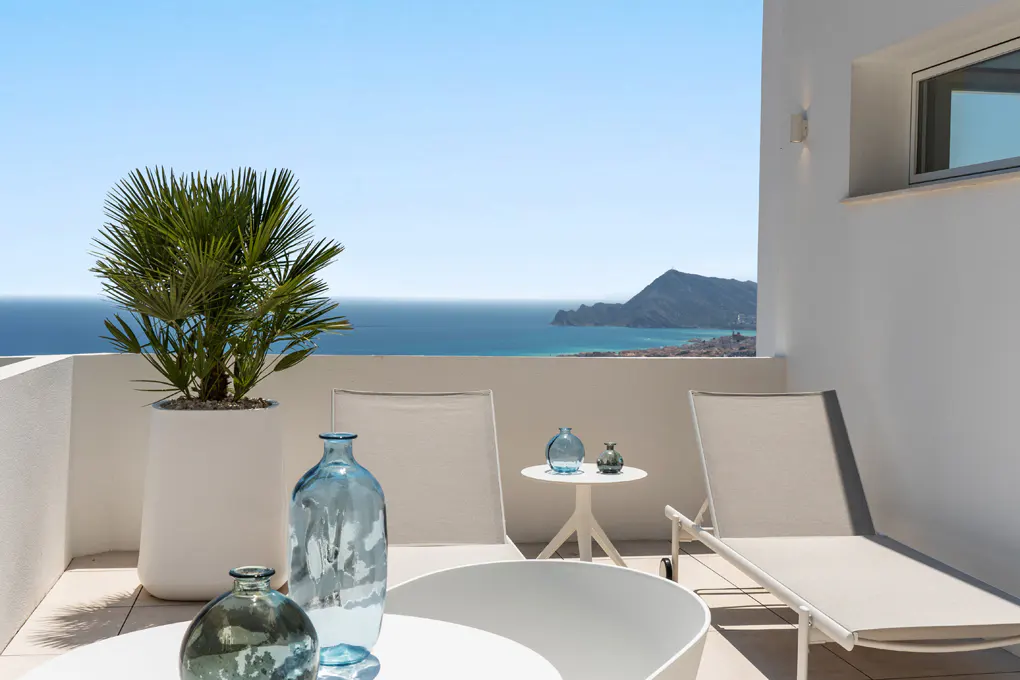 Balcony with ocean view. White lounge chairs, tables, and a potted palm tree. Blue glass vases add color. Bright, sunny day.