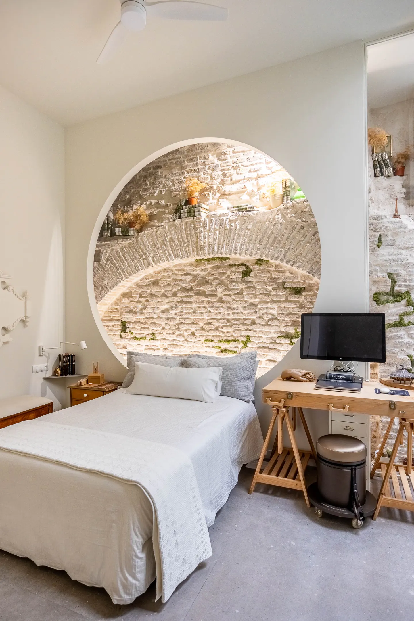 Bedroom with white walls, bed, and desk. An oval window shows a stone wall with plants. A ceiling fan is visible.