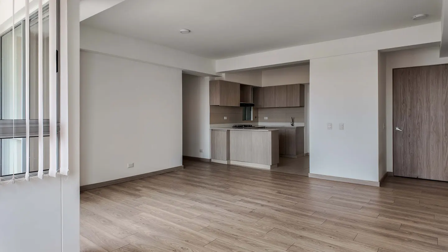 Bright, empty apartment with wood floors, white walls, and an open kitchen with light brown cabinets. Window with blinds on the left.