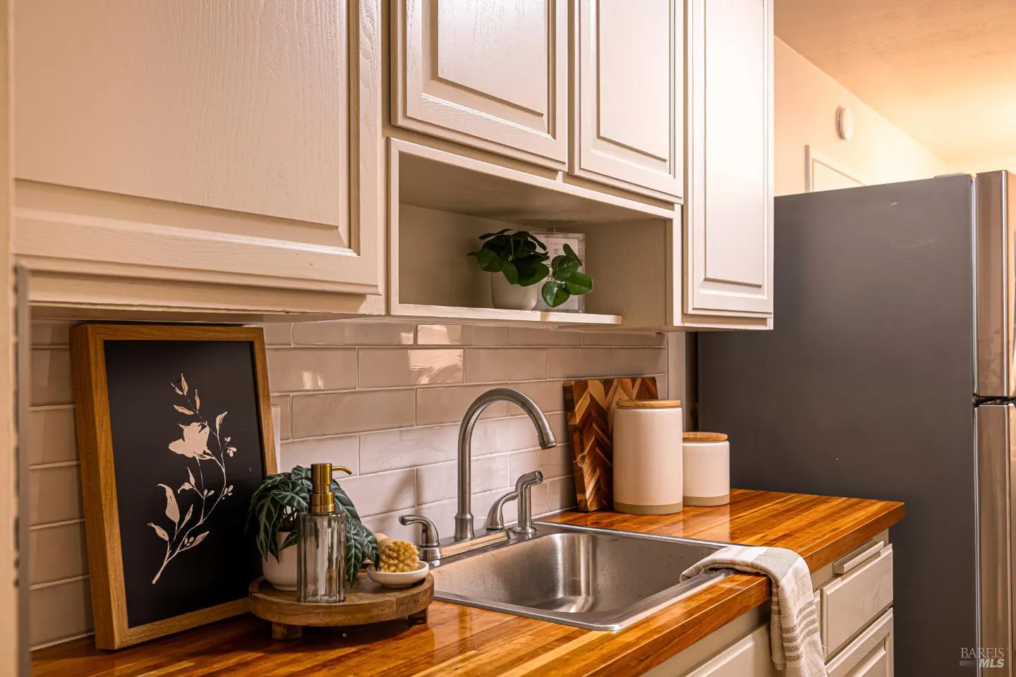 A kitchen view features a stainless steel sink, wood countertop, and cream-colored cabinets. A framed floral print and plants add a decorative touch.