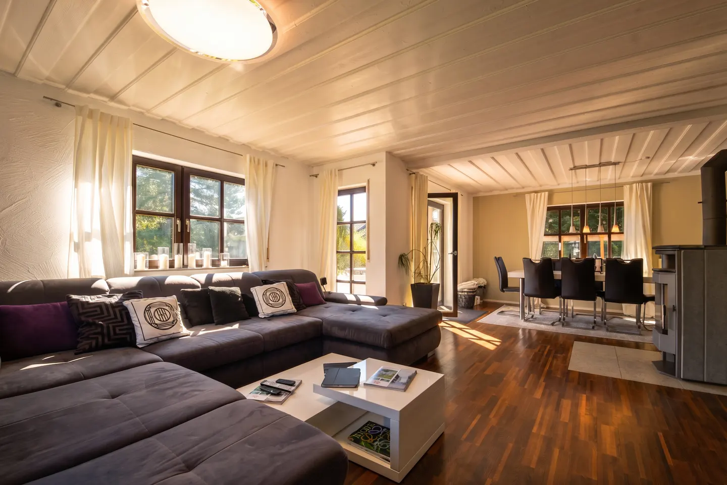 Bright living room with a gray sectional sofa, white coffee table, and wood floors. Dining table with black chairs in the background.