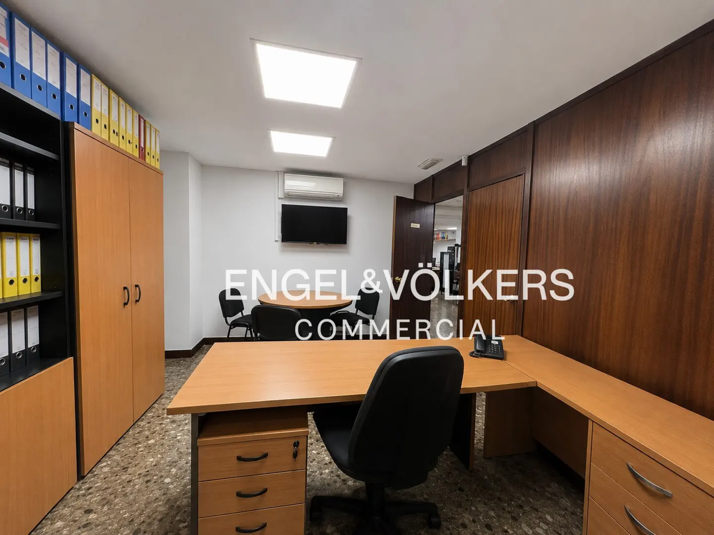 Office interior with a desk, black chair, and filing cabinets. A round table and chairs are in the background. The Engel & Völkers logo is superimposed.