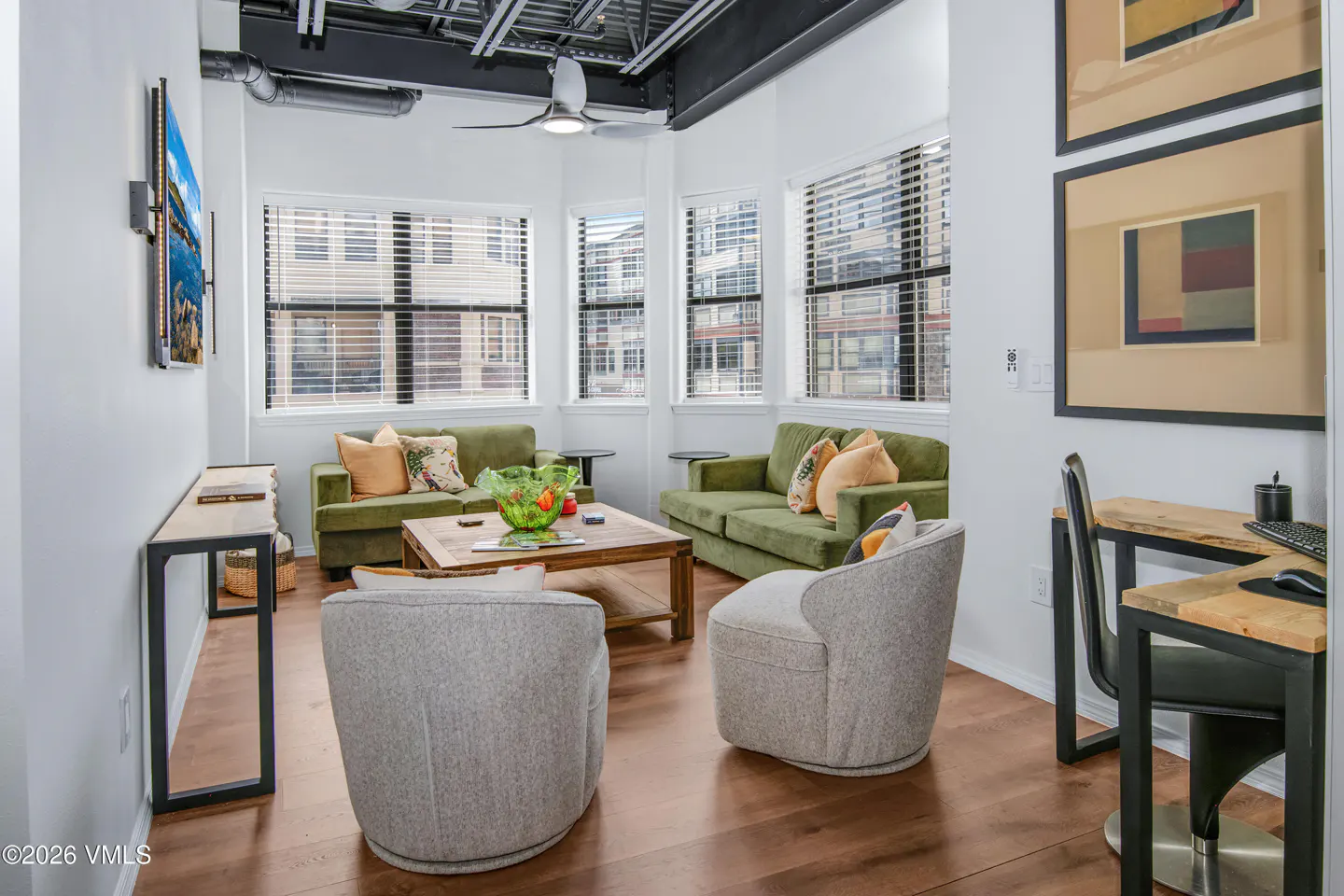 Bright living room with wood floors, white walls, and exposed ceiling. Two green sofas face two gray chairs around a wood coffee table. A desk sits to the right.