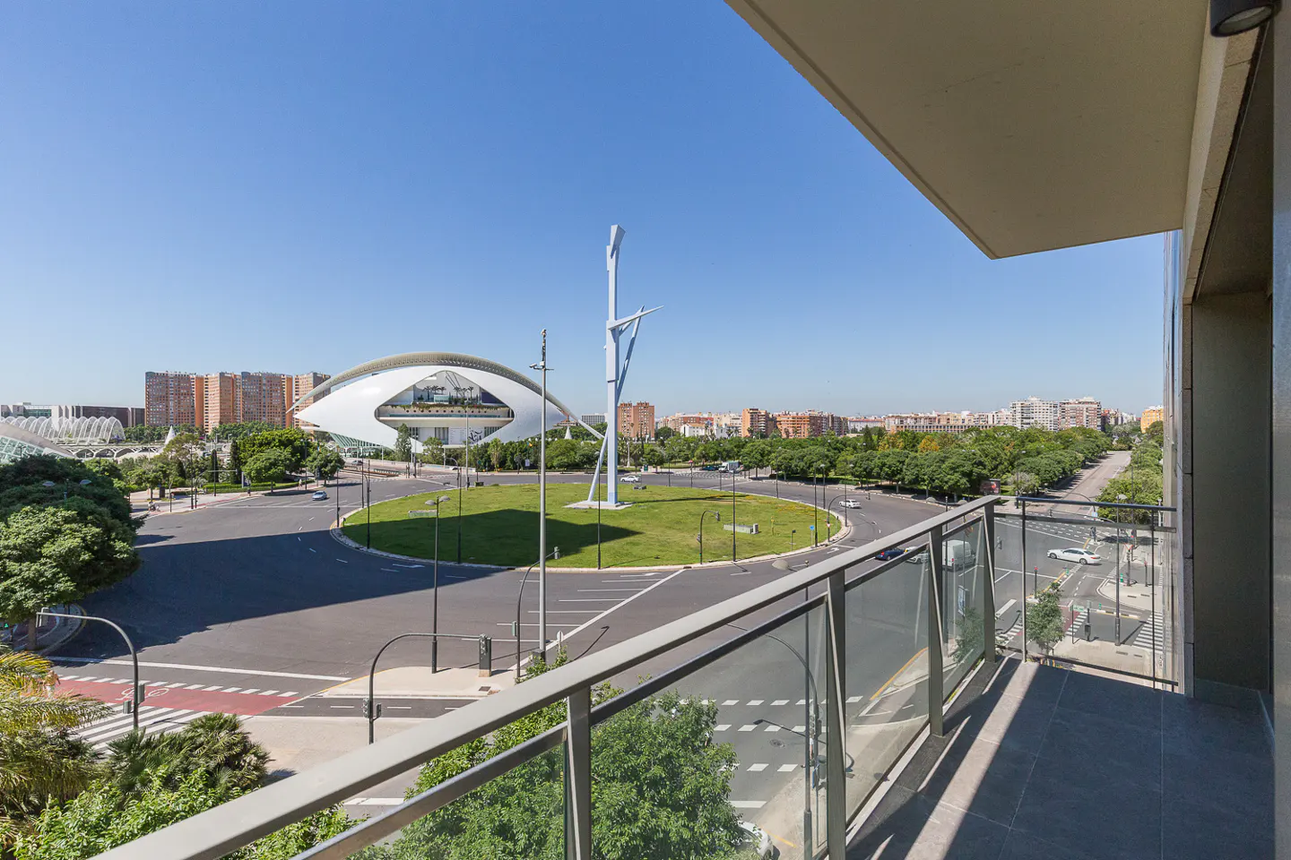 View from a balcony with glass railings overlooking a city with modern architecture and a roundabout.