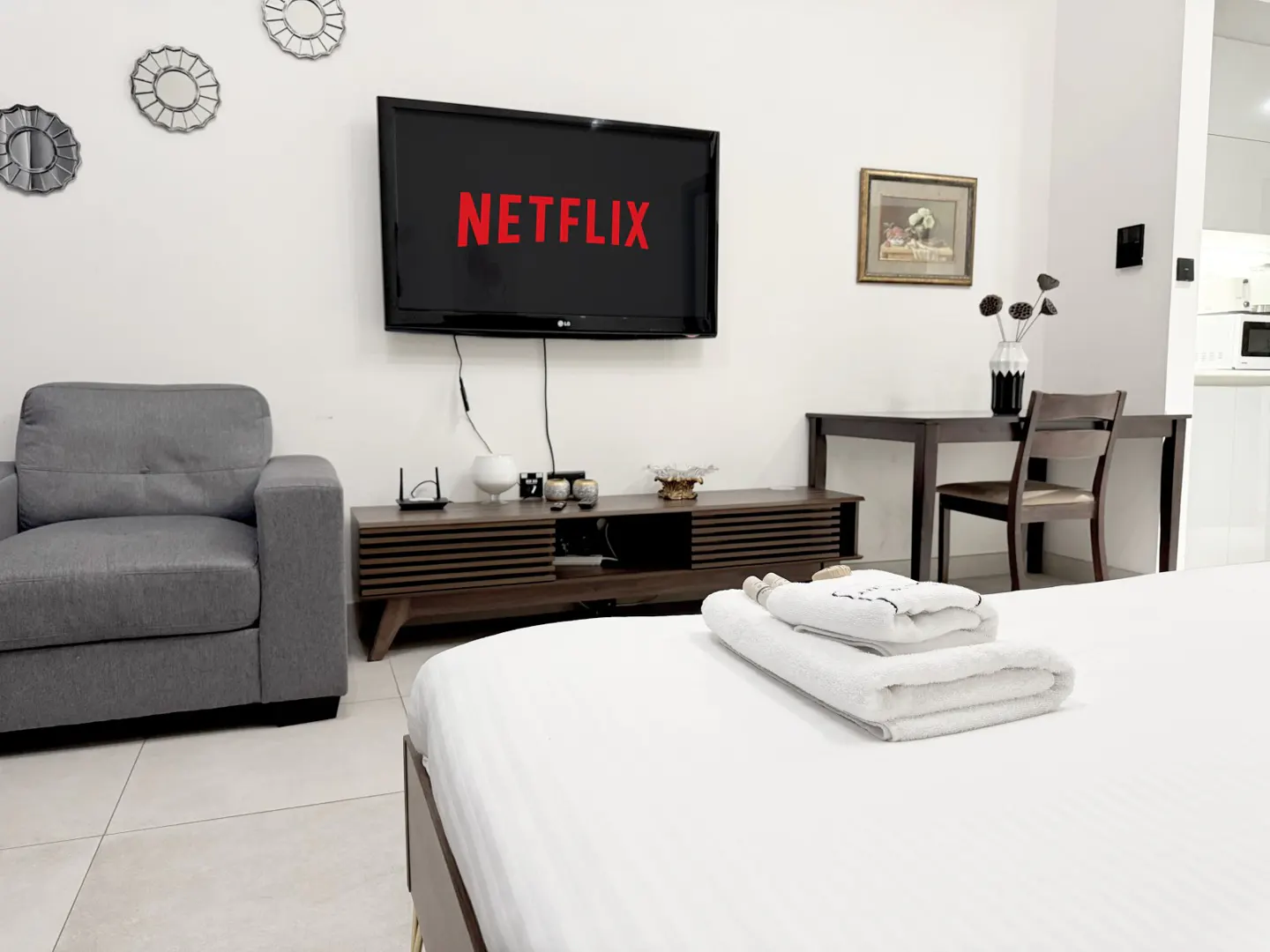 Bright studio apartment with a gray armchair, a TV with Netflix on, and a bed with folded white towels.