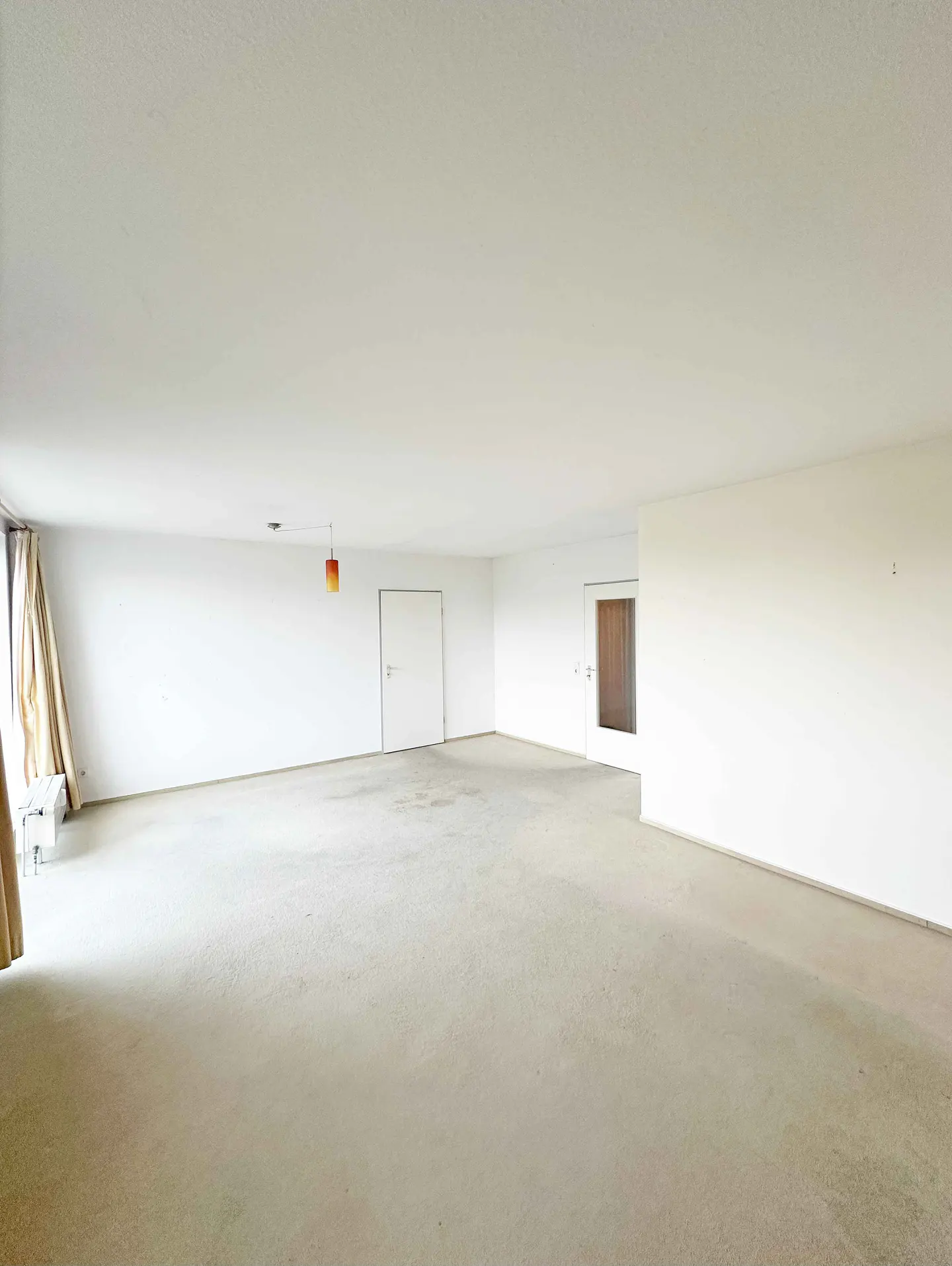 Spacious, empty room with white walls, beige carpet, two white doors, and a single hanging orange pendant light.