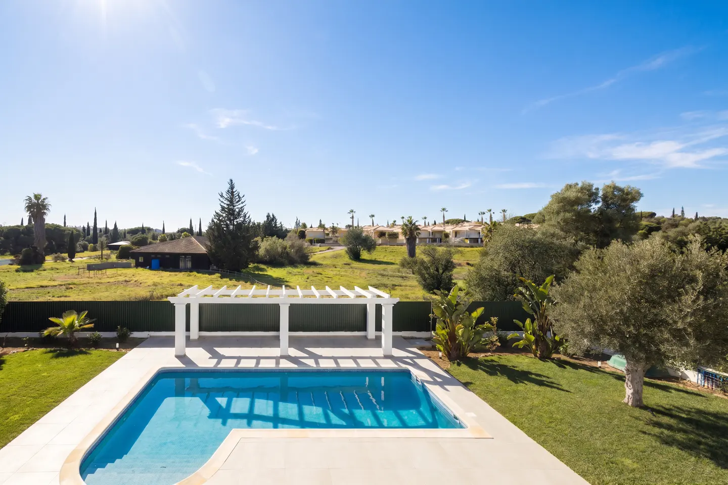 L-shaped swimming pool with a white pergola on a patio, surrounded by a green lawn and trees under a clear blue sky.