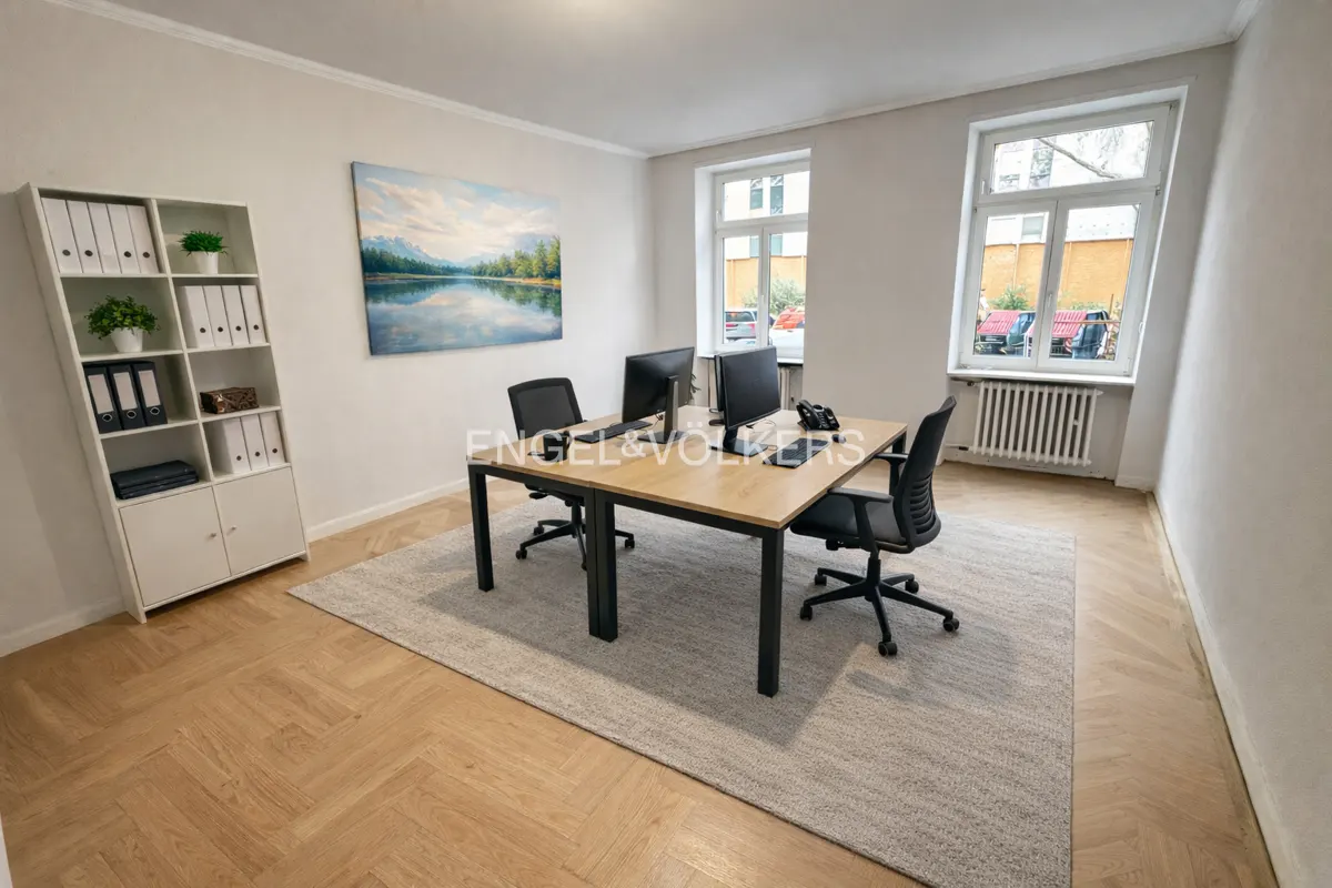 Office space with two desks, black chairs, computers, and a white bookshelf. A landscape painting hangs on the wall. Windows overlook a street.