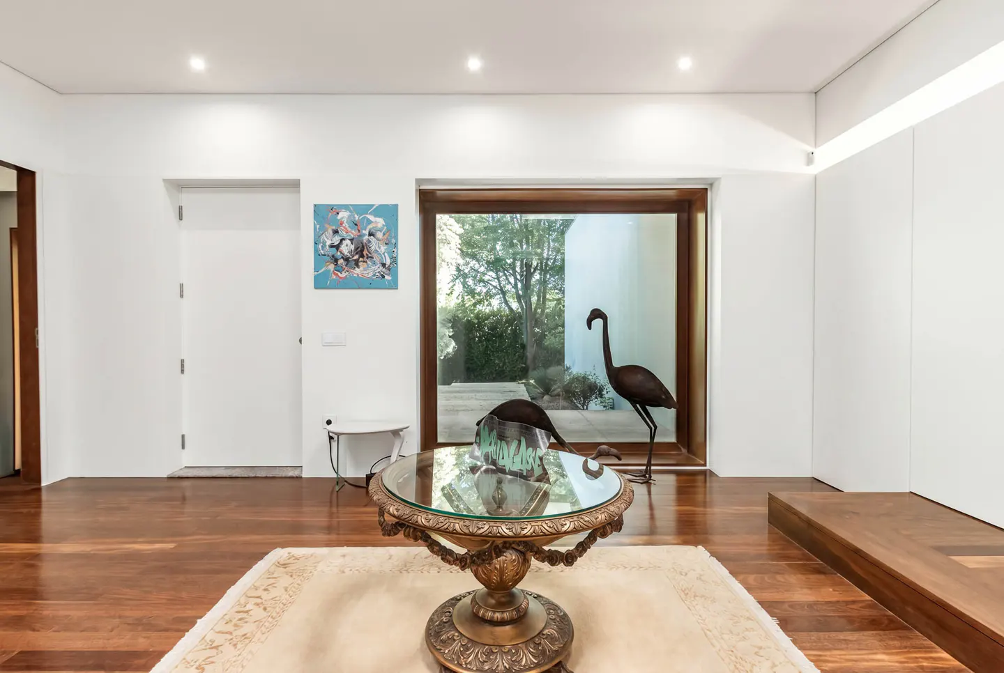 Bright living room with hardwood floors, white walls, and recessed lighting. A glass-topped table sits on a rug, with flamingo statues nearby.