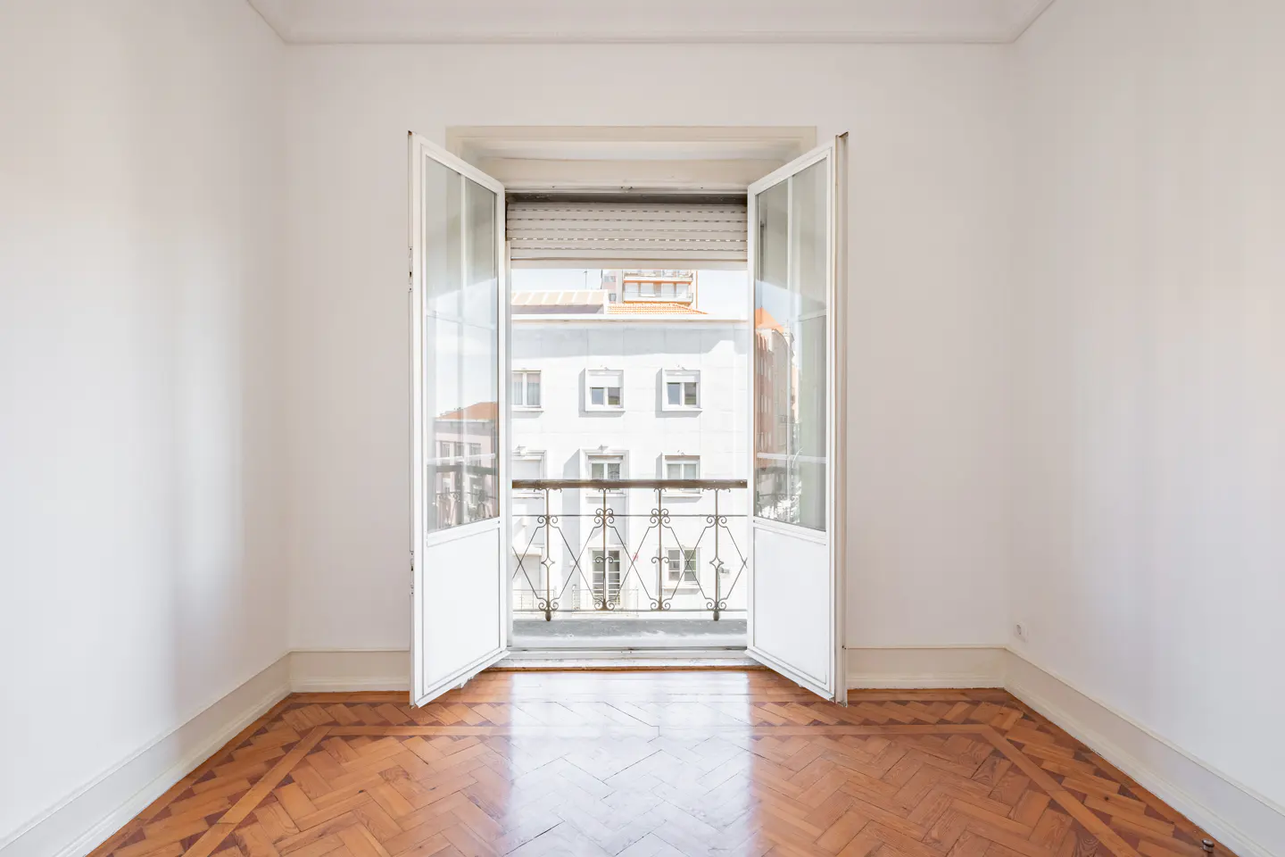 Empty room with white walls, polished herringbone wood floors, and open French doors leading to a small balcony.