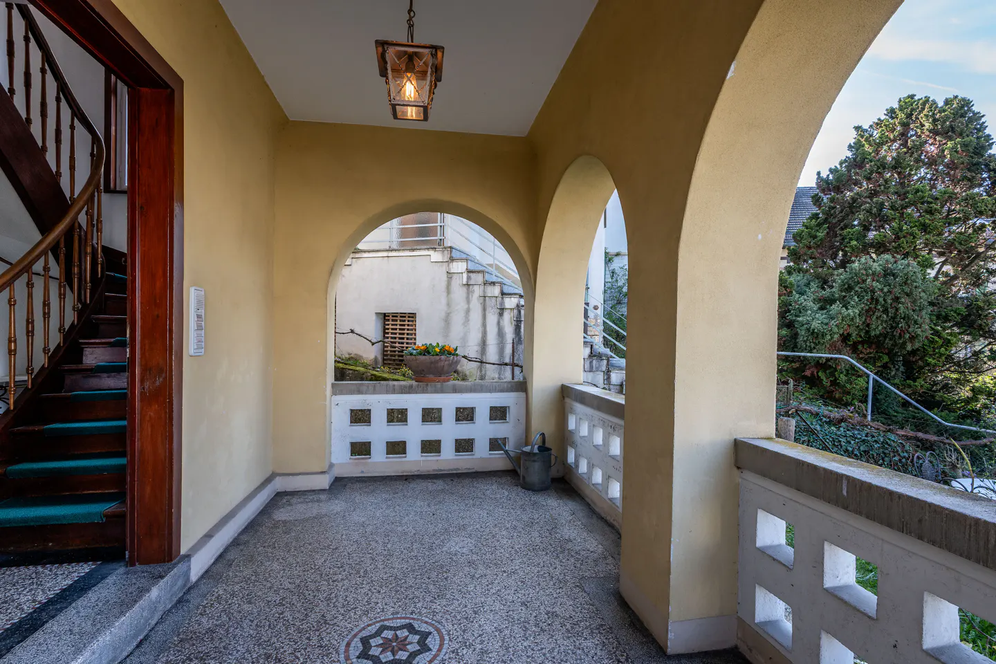 Arched entryway with yellow walls, a mosaic floor, and a wooden staircase. A hanging lantern illuminates the space. Balconies and greenery are visible outside.
