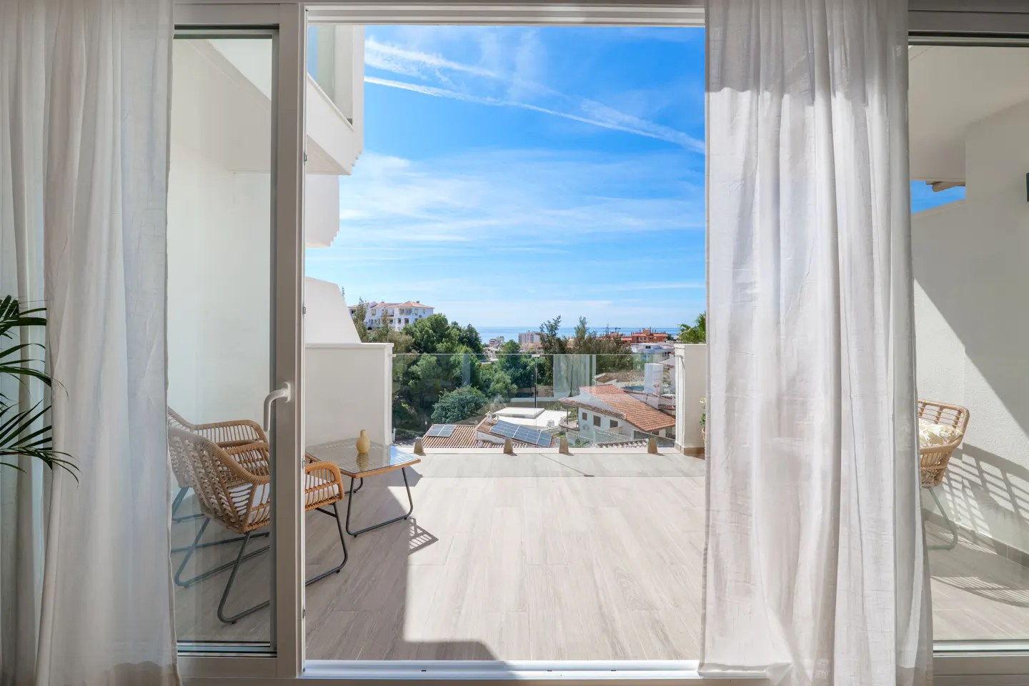 View from open patio doors onto a balcony with wicker chairs and a table, overlooking a sunny landscape with blue sky.
