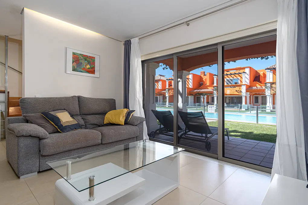 Living room with gray sofa, glass table, and sliding doors to a patio with pool and orange buildings.