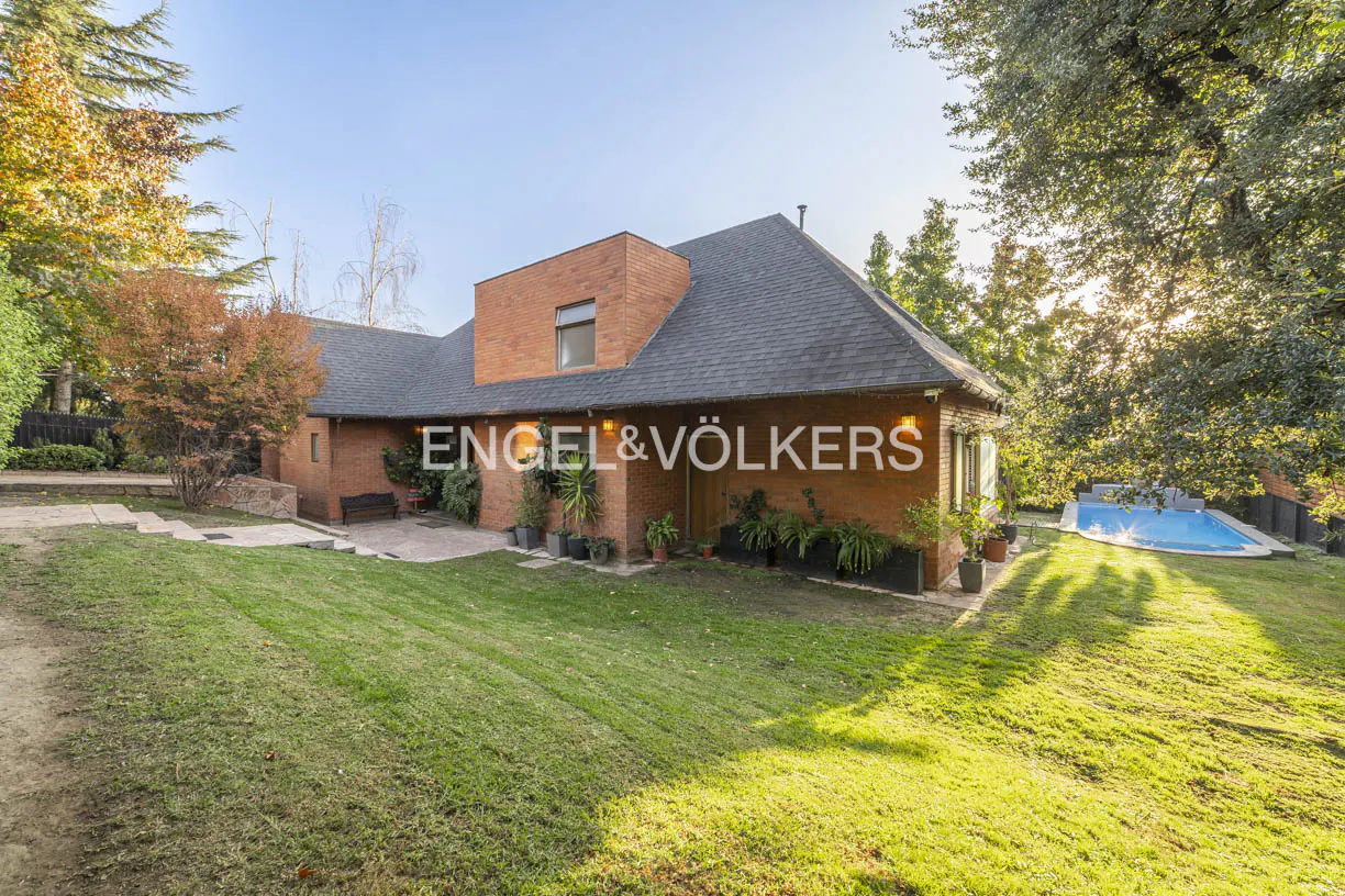 Exterior view of a brick house with a gray roof, green lawn, and a blue swimming pool in the background. "Engel & Völkers" logo on the house.