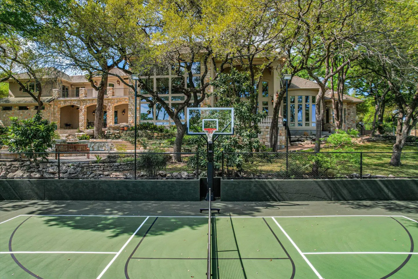 Outdoor basketball court with green surface and white lines, net, and hoop, in front of a large stone house and trees.