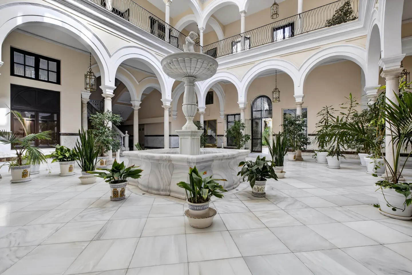 Interior courtyard with a marble fountain, potted plants, and arched walkways. Cream-colored walls and a tiled floor add to the elegant design.