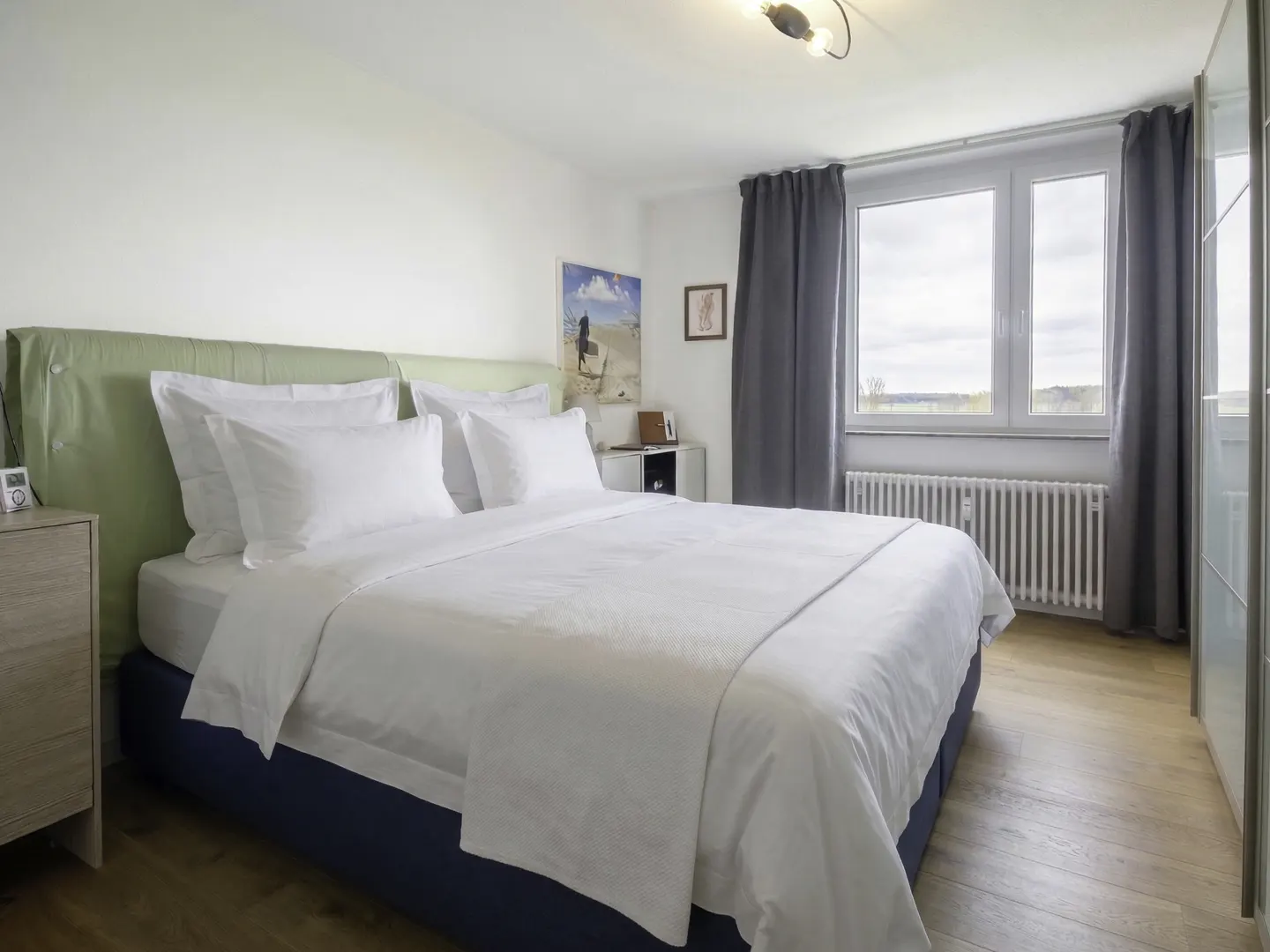 Bedroom with a large bed, white linens, and a light green headboard. Gray curtains frame a window with a view. Wooden floors and white walls.
