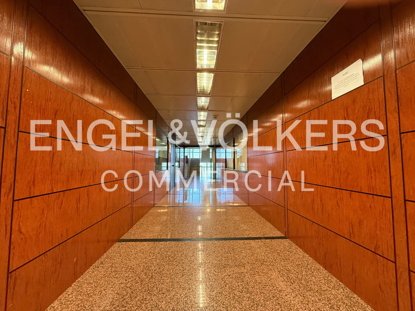 Hallway view of Engel & Volkers Commercial office with wood paneled walls and speckled floor. Bright lights illuminate the space.